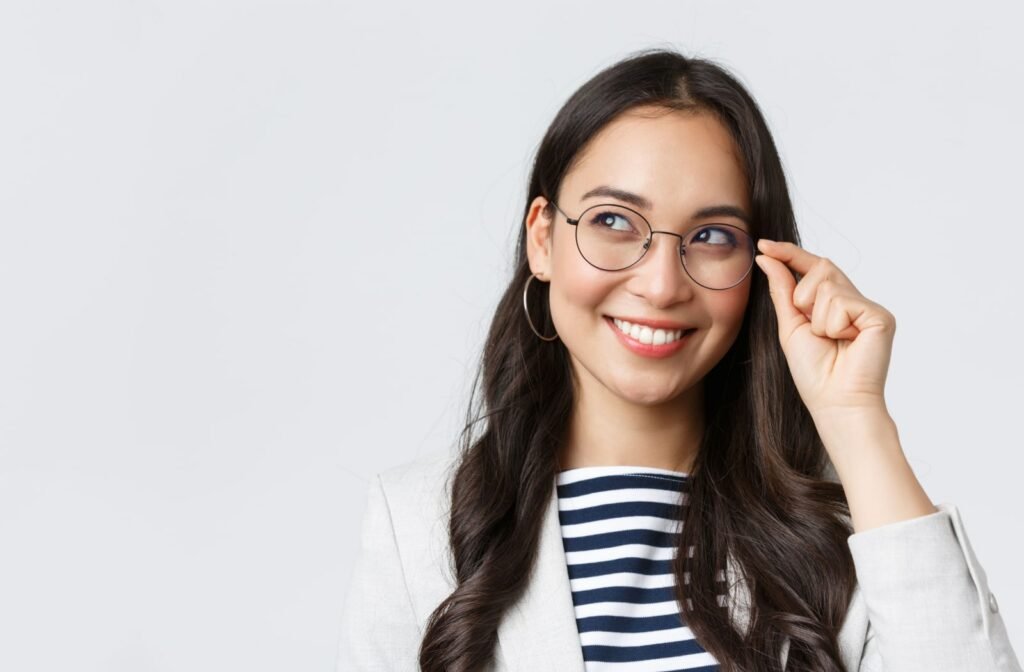 A woman smiling, looking up and touching the side of her glasses with her left hand