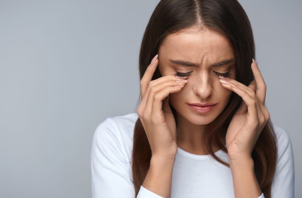 A woman wearing a white shirt in front of a grey background touching her eye's in discomfort as she is getting some pain from her contact lenses