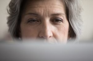 An older woman looking straight ahead at a computer screen, with the screen taking up only a small portion of the bottom of the image.